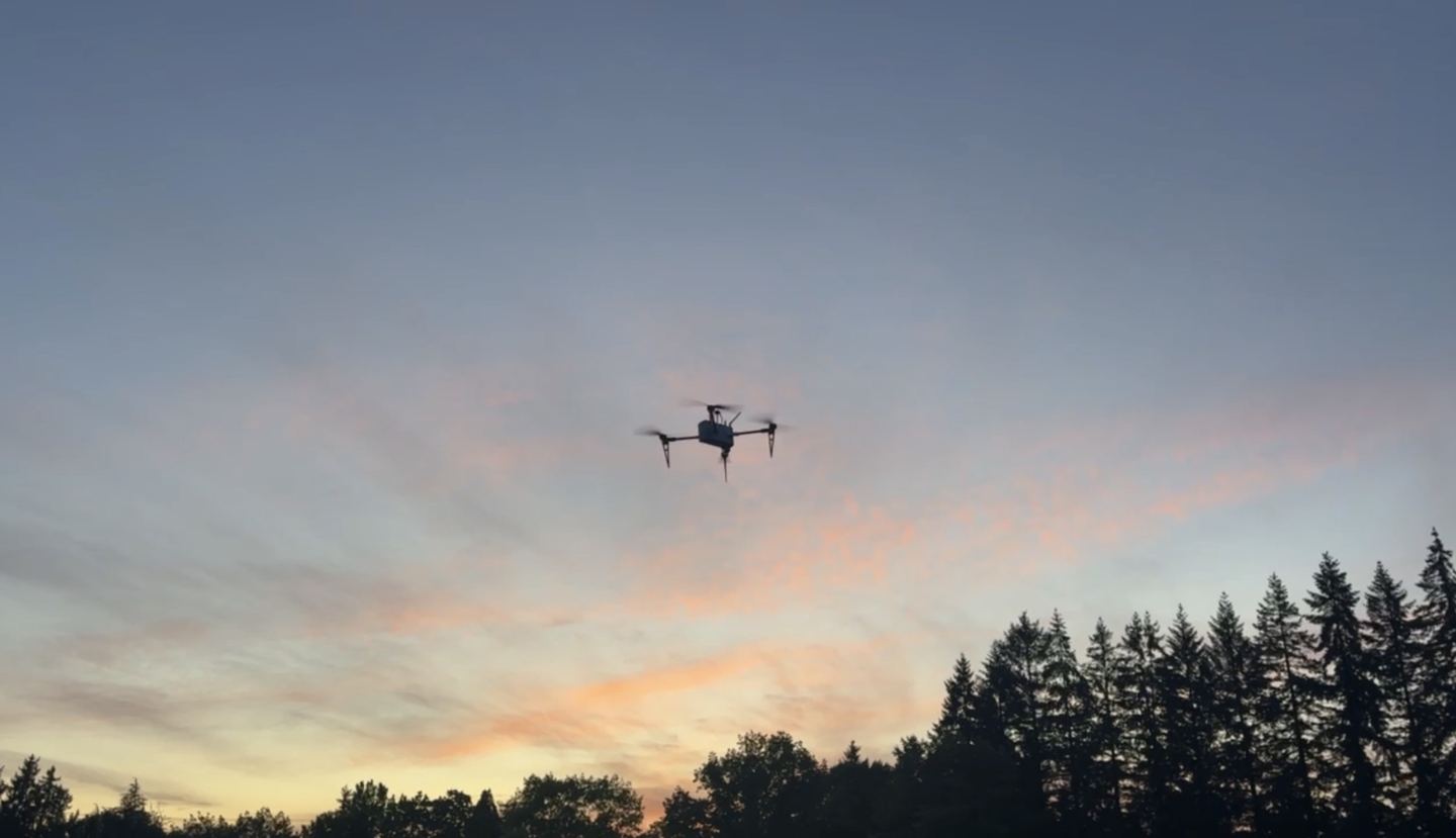 The Aegis quadcopter flying at sunset above a treeline, silhouetted against pink and orange clouds